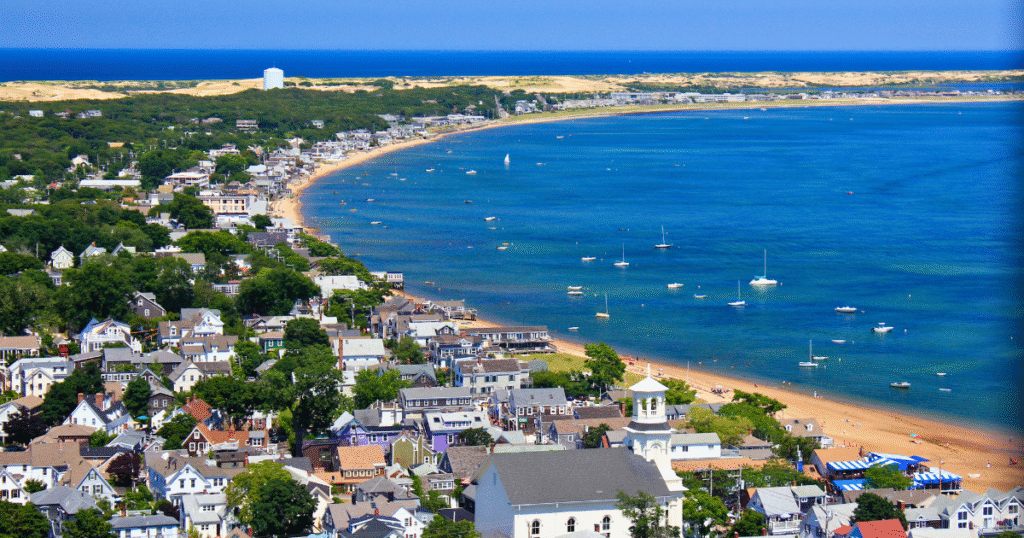 Aerial view of a Cape Cod beach town with boats in the harbor, colorful homes, and summer shoreline.